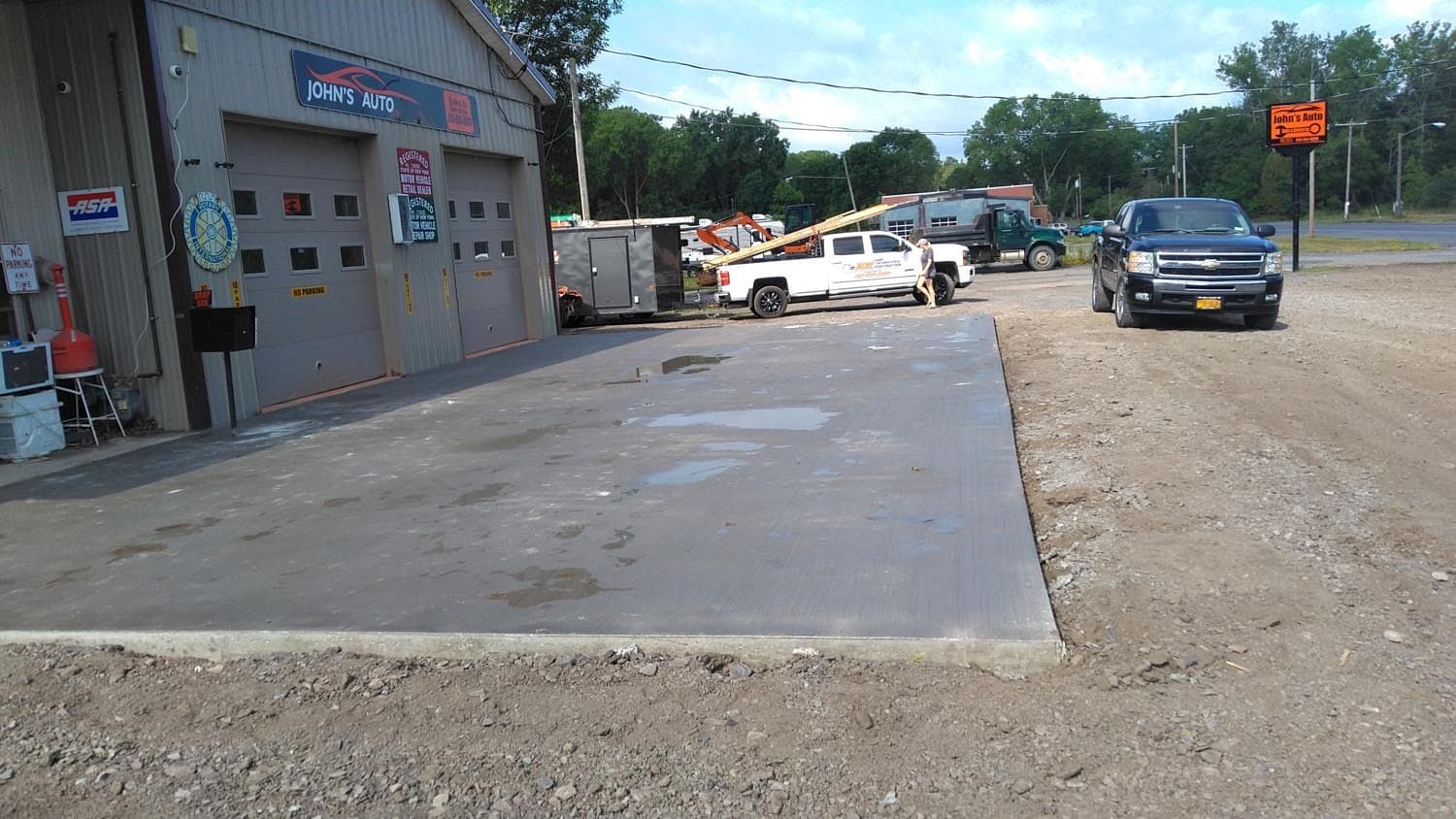 Fresh concrete driveway at John's Auto shop with vehicles and construction equipment