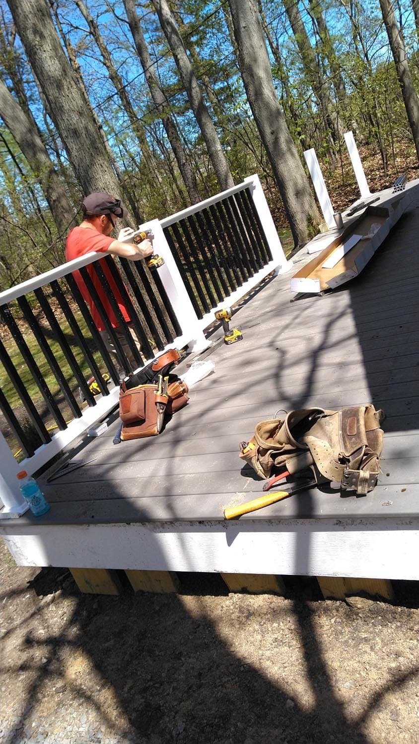 Worker installing white and black railing on outdoor deck surrounded by trees