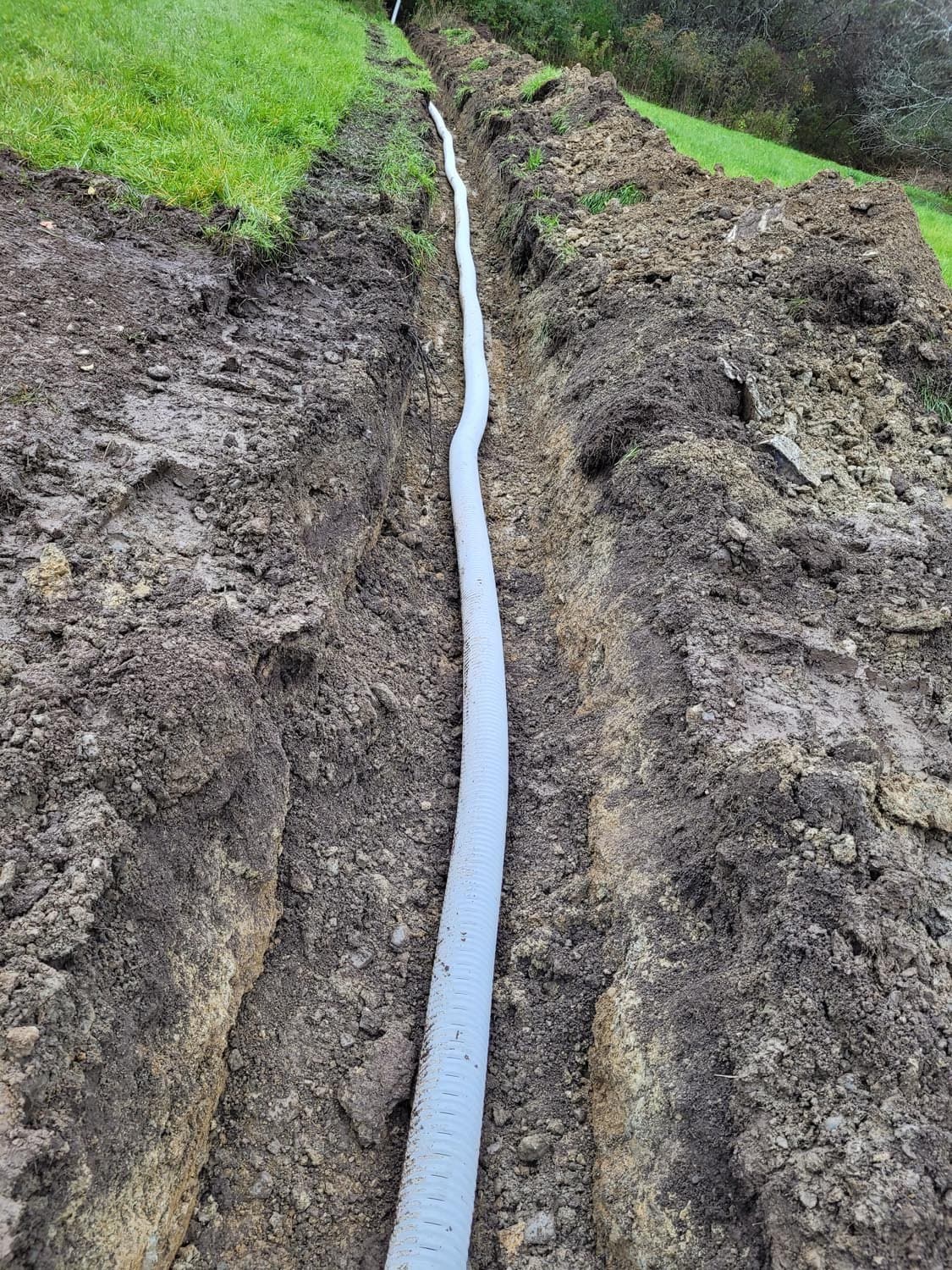 White drainage pipe installed in excavated trench with dirt mounds on sides