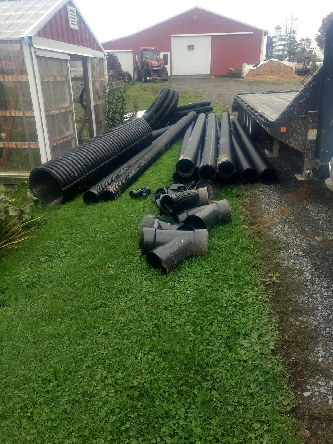 Black drainage pipes and fittings on grass at farm worksite with red barn