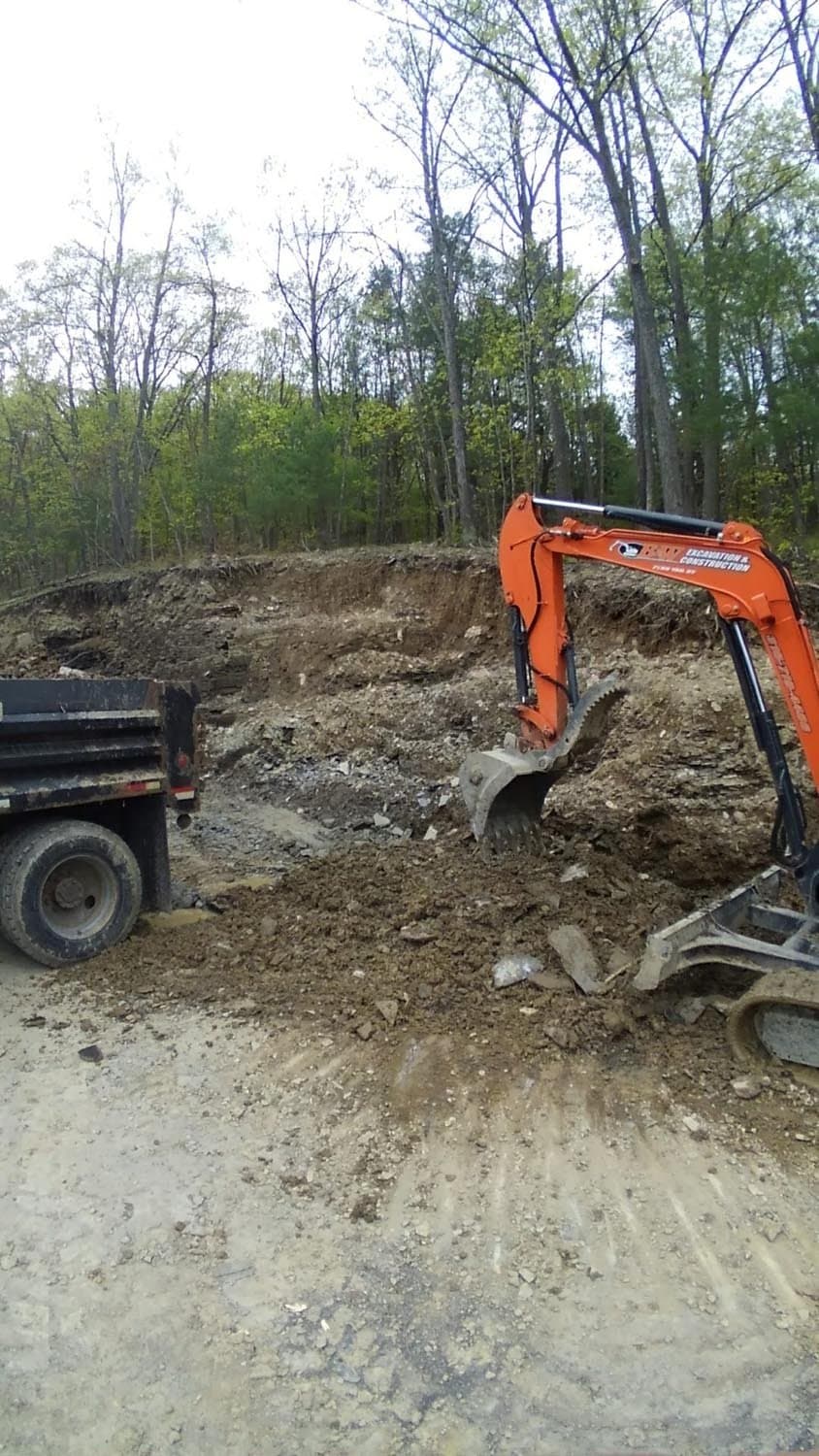 Orange excavator digging foundation with dump truck at wooded construction site