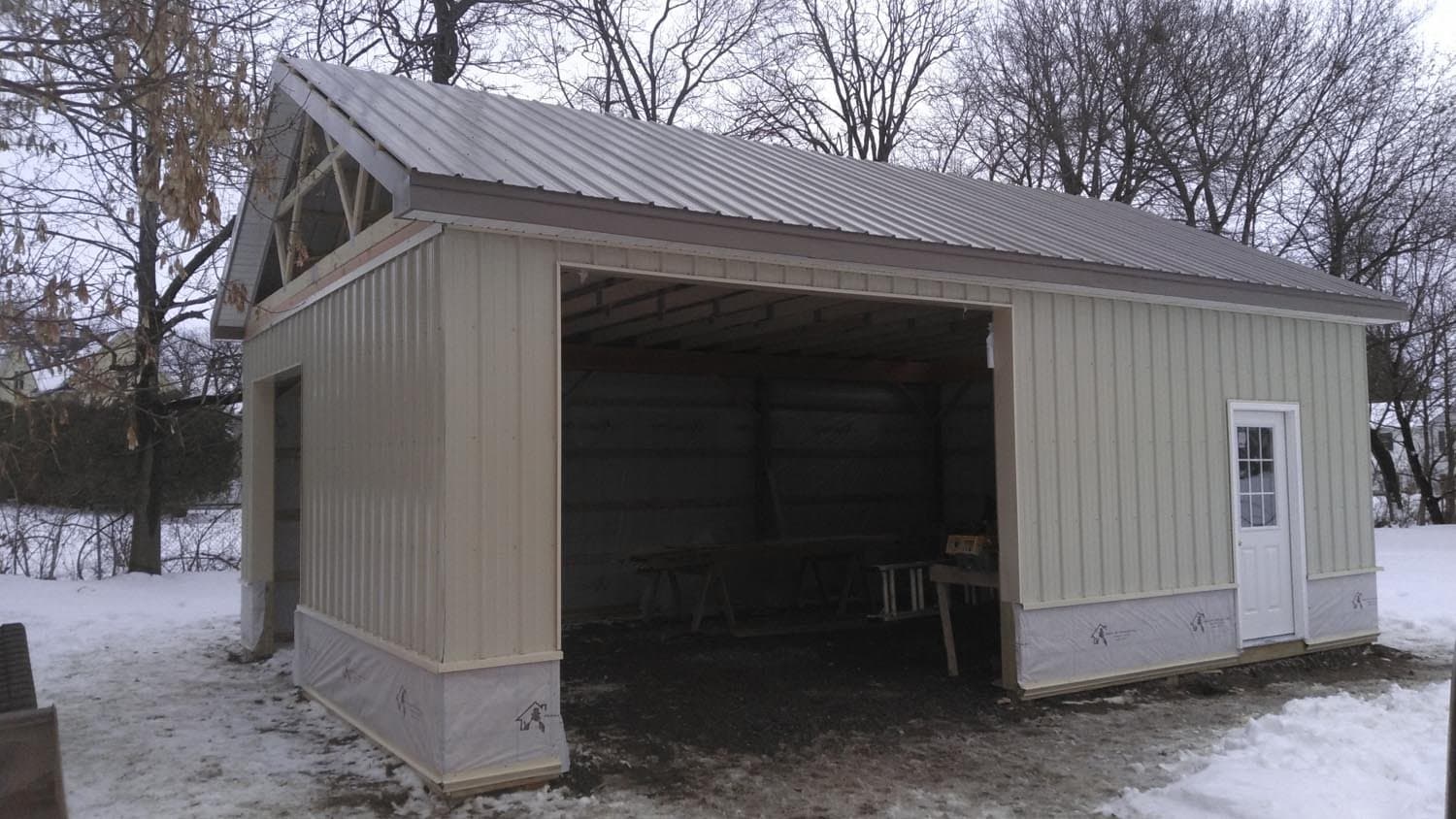 Beige metal pole barn with open garage bay and white entry door in winter