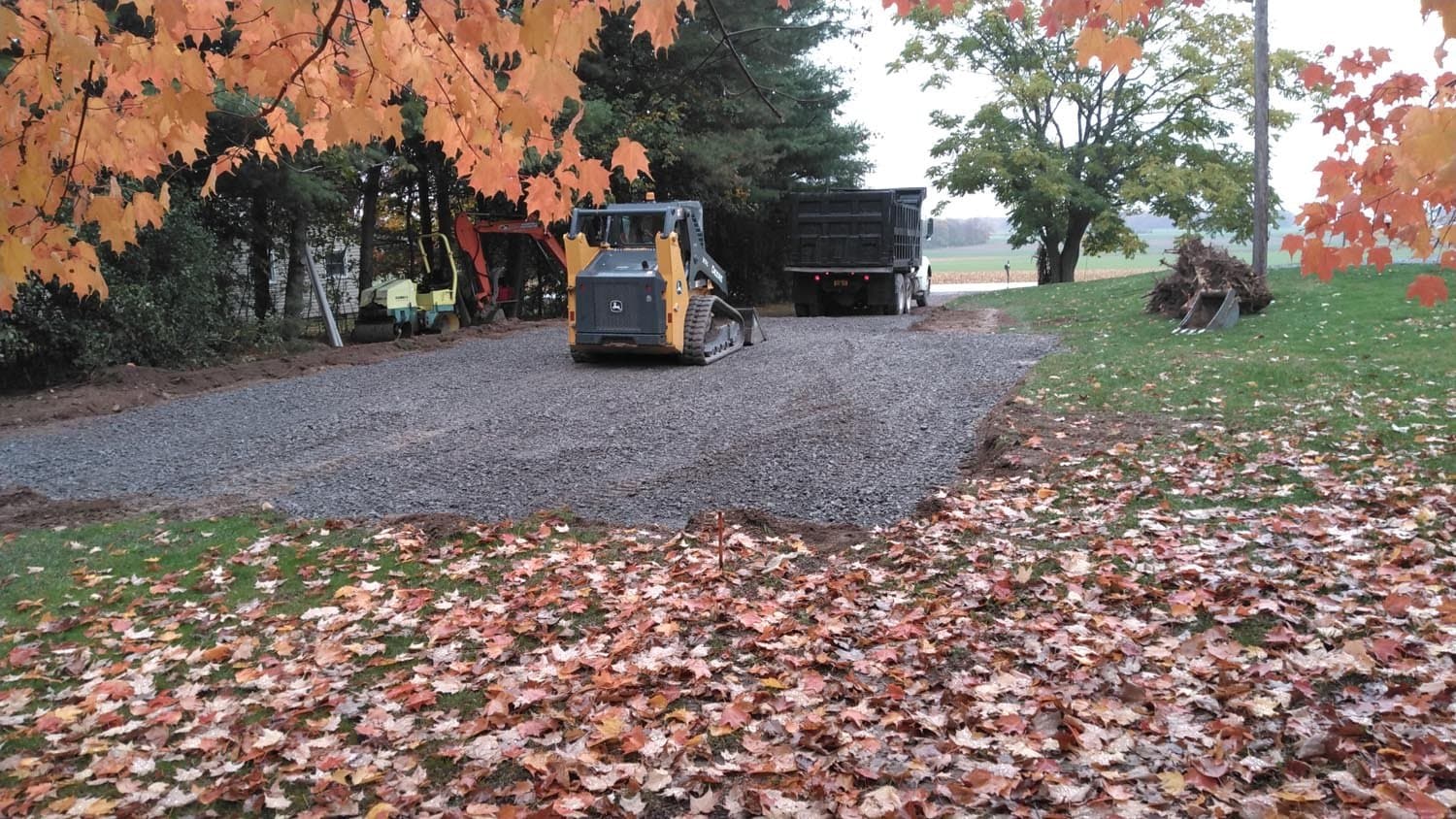 Compact track loader and dump truck working on new gravel driveway in autumn