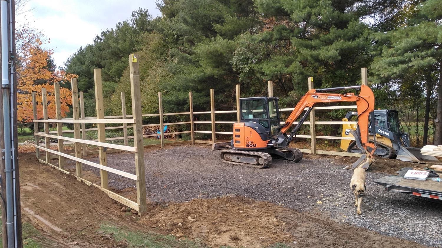 Wooden pole barn frame under construction with orange excavator and yellow skid steer on gravel base