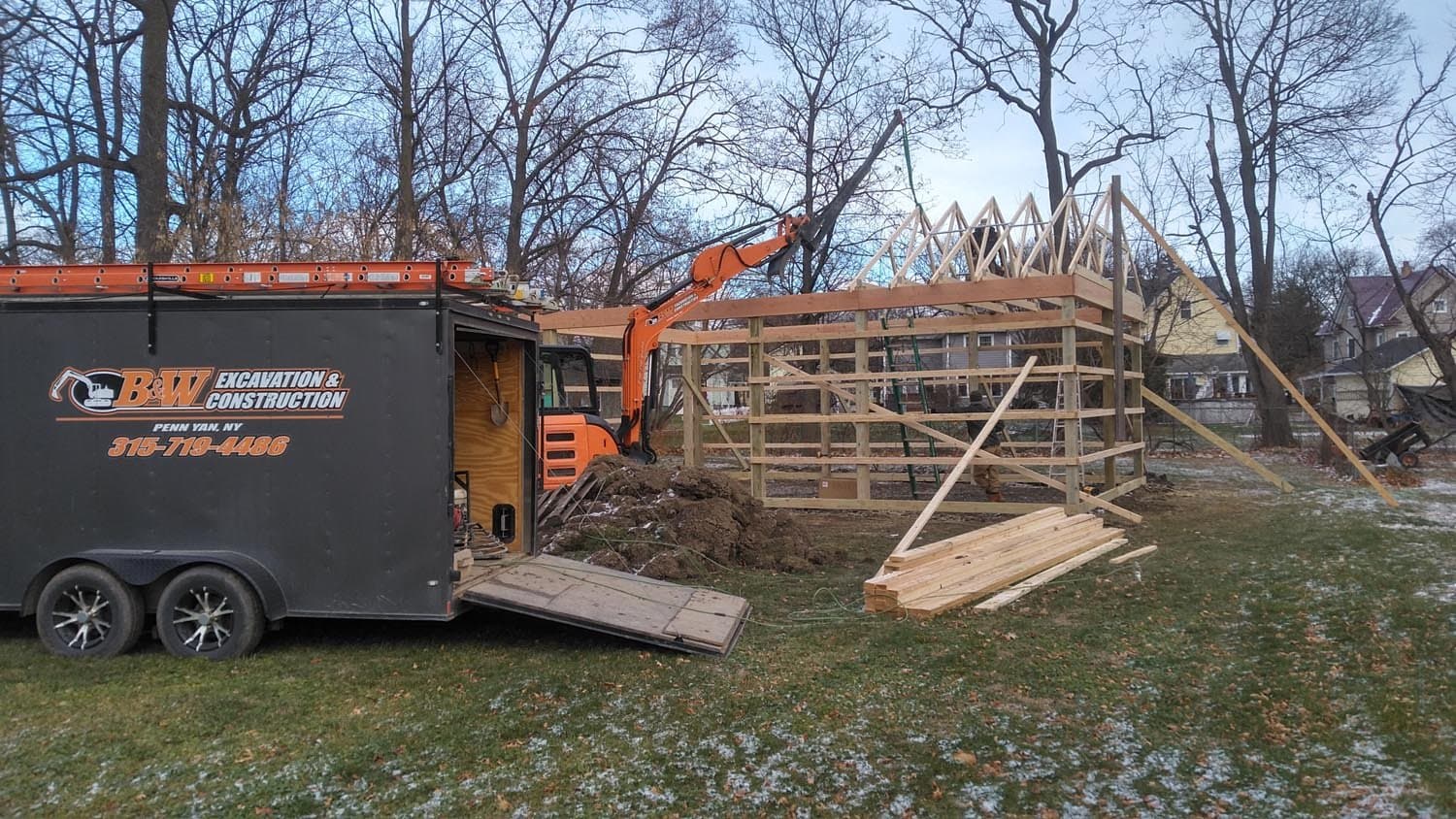 Wooden pole barn frame under construction with B&W Excavation trailer and orange excavator on site