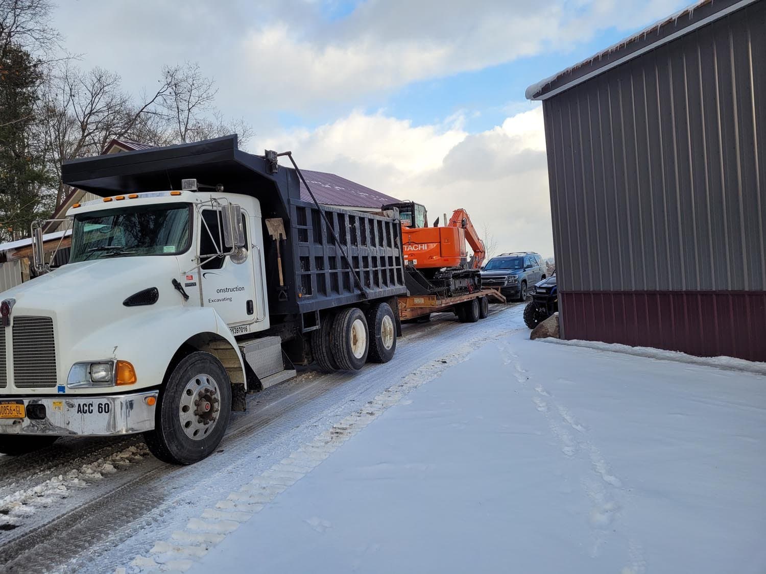 White dump truck and orange excavator on snowy site next to metal pole barn