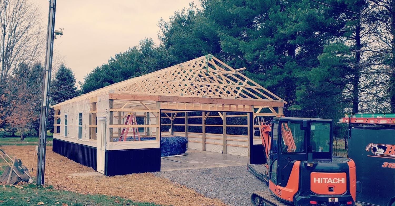 Wooden frame of pole barn under construction with exposed roof trusses and Hitachi excavator