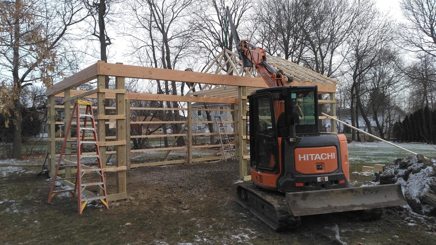 Pole barn timber frame under construction with excavator and ladder on site