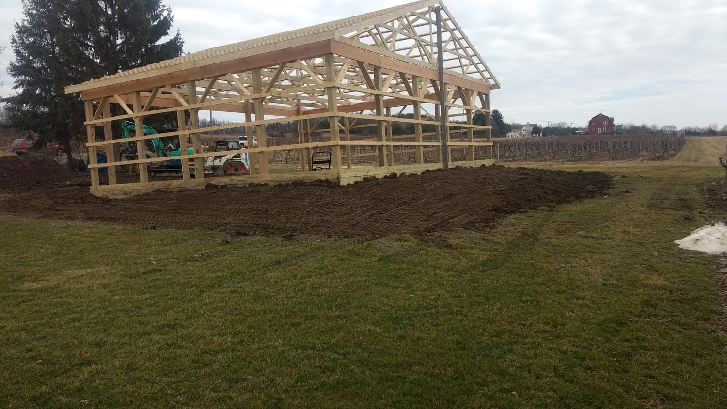 Wooden pole barn frame under construction in open field with vineyard and red barn in background