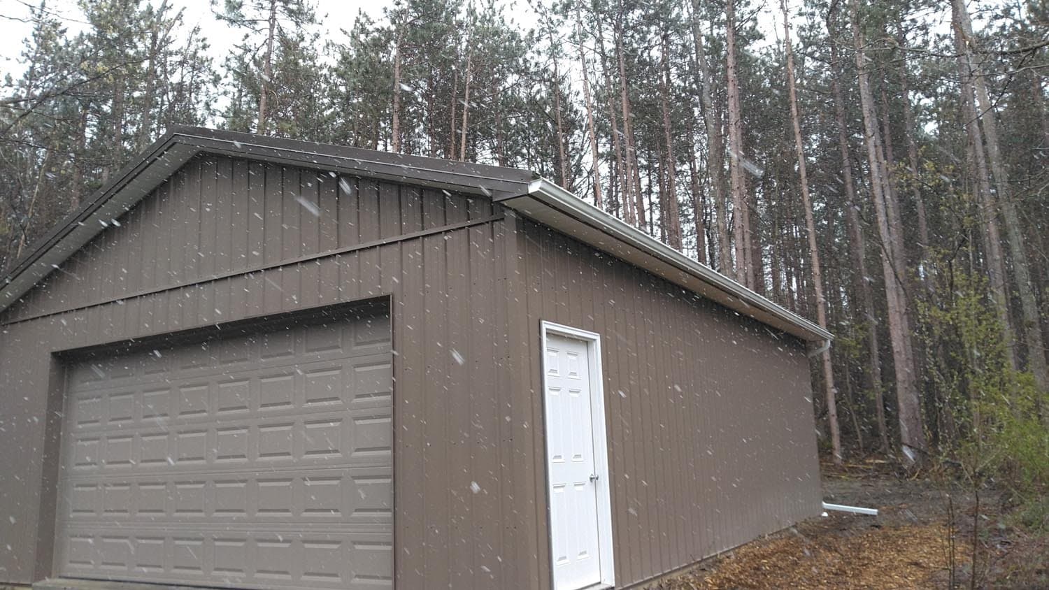 Brown pole barn with garage door and white entry door during snowfall