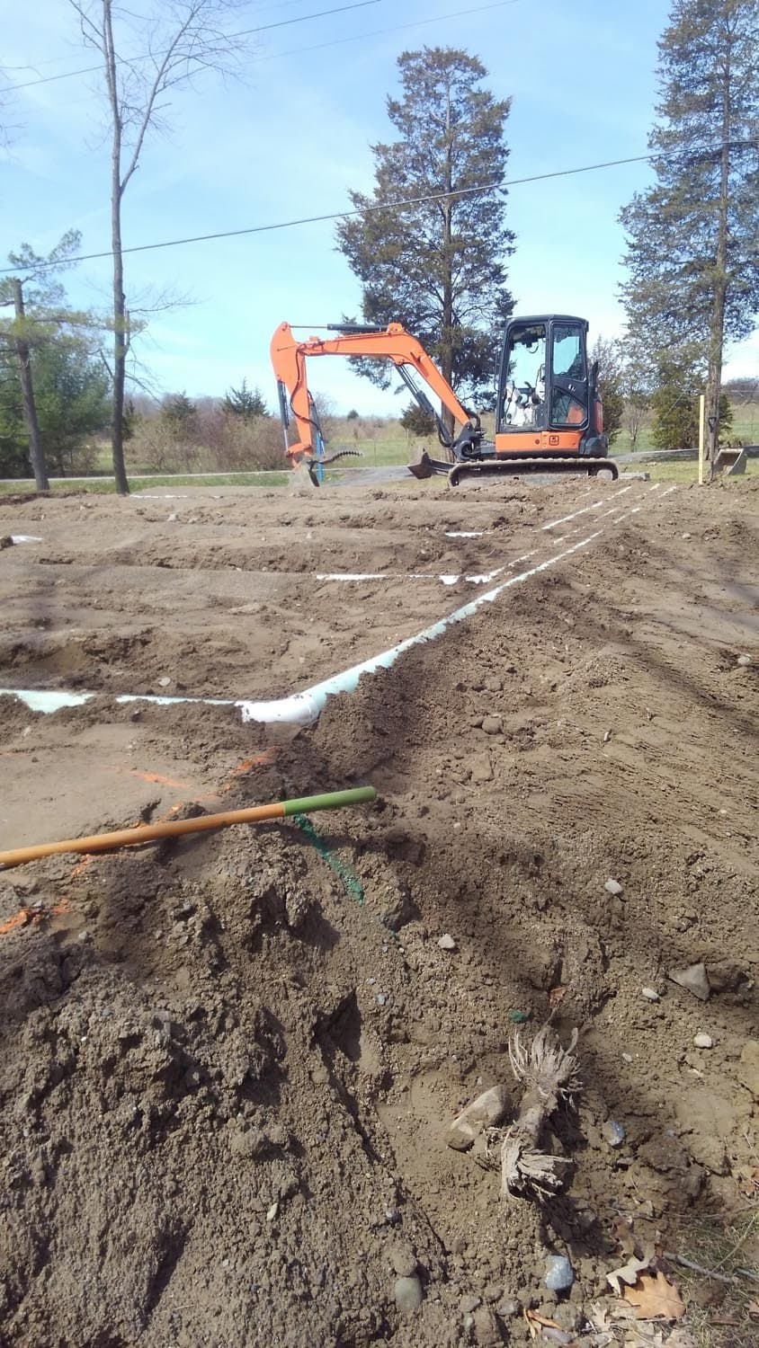 Orange excavator digging septic system foundation with white marking lines and exposed utility pipes