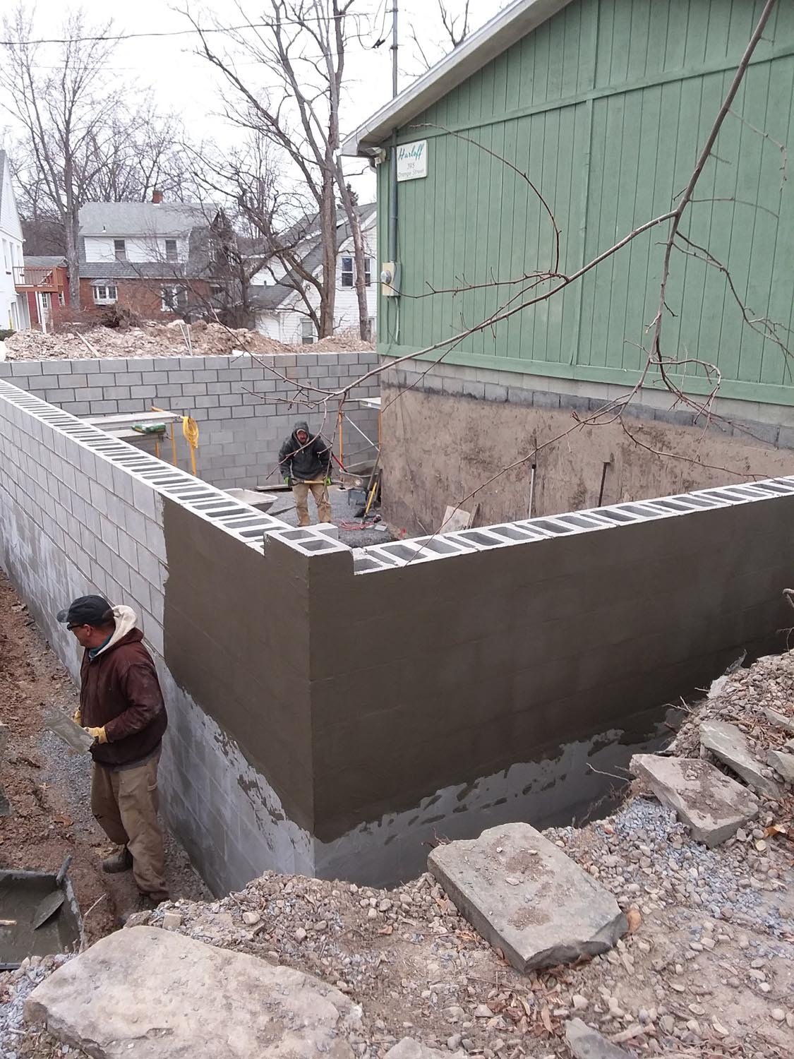 New concrete-block foundation wall with dark waterproofing and a worker in the excavation beside a green building