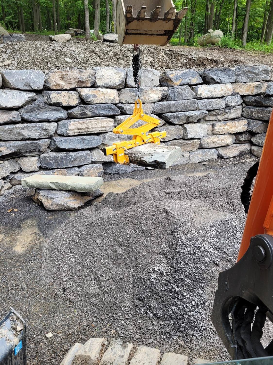 Excavator using a yellow clamp to lower a large stone onto a natural-stone retaining wall, with crushed stone at its base